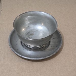 May include: A vintage metal teacup and saucer. The cup has a round bowl shape with a pedestal base, and the saucer is a matching round shape. Both items have a dull, silver-grey finish, possibly pewter. The set is likely antique and would be measured in centimetres.