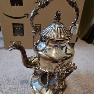 Puede incluir: Tetera plateada ornamentada con un asa y un pico decorativos. La tetera se encuentra sobre un soporte a juego con detalles intrincados. El diseño presenta una tapa acanalada y un remate floral, mostrando una estética vintage.