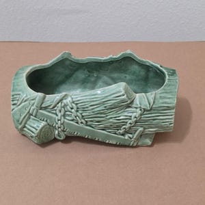 Pode incluir: Um vaso de cerâmica verde em forma de tronco, com uma abertura retangular. O vaso tem entalhes detalhados de grão de madeira, marcas de serra e uma corrente. A cor é verde suave e o design é rústico.