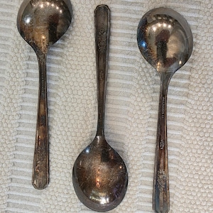 May include: Three antique silver serving spoons. The spoons have ornate detailing on the handles and large, rounded bowls. The spoons are arranged on a textured, off-white surface.