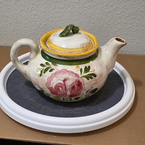 May include: Cream-colored ceramic teapot with a floral design. The teapot features a large pink rose, green leaves, and a yellow and green rim. It has a curved handle and a spout. The lid has a green handle. The teapot sits on a white and gray tray.