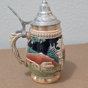 May include: A decorative beer stein with a detailed landscape scene. The ceramic mug features a silver metal lid and handle. The design includes a red-roofed building, trees, and a water wheel, with a cream-colored base and handle.