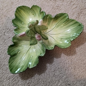 May include: A decorative ceramic serving dish in the shape of three overlapping green leaves. The dish features a central floral arrangement with pink and white accents. The leaves have a glossy finish and detailed veining.