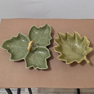 May include: Two ceramic leaf-shaped serving dishes. One is a large, three-lobed leaf with a yellow handle. The other is a smaller, single leaf with a textured surface and a distressed, yellow-brown rim. Both are shades of green.