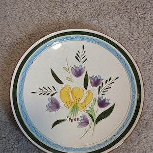 May include: Round ceramic plate with a floral design. The plate features a central yellow lily surrounded by purple flowers and green leaves. A green and blue border frames the white center.