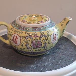 May include: A yellow ceramic teapot featuring a lid, a curved handle, and a spout. The body is decorated with a repeating scroll pattern, pink floral motifs, a geometric border, and two circular medallions containing red Chinese characters.