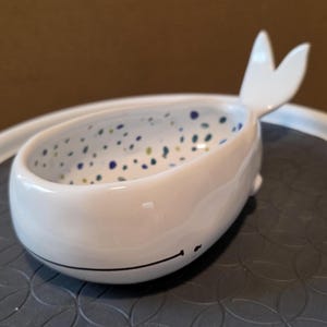 May include: A white ceramic whale-shaped bowl with a whimsical design. The bowl features a wide opening, a tail fin, and small side fins. The interior is speckled with blue and green dots, and a black line forms a smiling mouth.