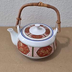 May include: A white ceramic teapot featuring a rounded body with a decorative pattern of red panels, gold floral motifs, and green accents. The teapot includes a matching lid and a curved, light brown woven bamboo handle attached to two loops.