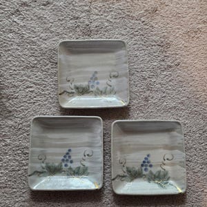 May include: Three square ceramic plates with a hand-painted grape design. Each plate features a light gray background with a watercolor effect, a cluster of blue grapes, and green leaves. The edges have gold accents.