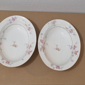 May include: Two oval-shaped white ceramic serving bowls featuring a delicate floral pattern with pink, yellow, and purple flowers along the rims and a thin gold-toned trim around the edges. The center of each bowl is plain white with a small floral motif.