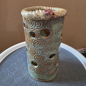 May include: A handmade ceramic vase with a cylindrical shape. The vase features a textured surface with swirling patterns and circular cutouts. The rim is adorned with a braided design and small, pinkish-red accents. The vase has a green and brown color palette.