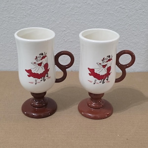 Peut inclure: Deux tasses en céramique de couleur crème avec des bases sur pied marron et des anses circulaires marron assorties. Chaque tasse présente une illustration graphique d'un couple dansant en tenue rouge et noire, avec une jupe rouge.