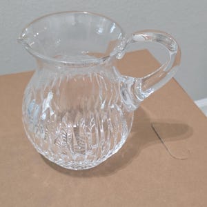 Könnte beinhalten: Ein Krug aus klarem Glas mit rundem Körper und Henkel. Der Krug hat ein strukturiertes Schliffmuster und einen Ausguss. Der Krug steht auf einer braunen Oberfläche.