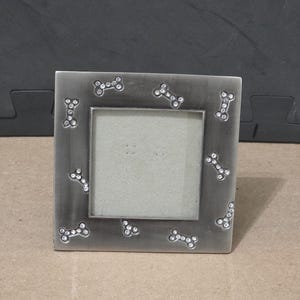 May include: A square, brushed silver-tone metal picture frame featuring a repeating pattern of small, rhinestone-encrusted dog bone shapes scattered across the border. The frame has a central square opening for a photograph and a smooth finish.