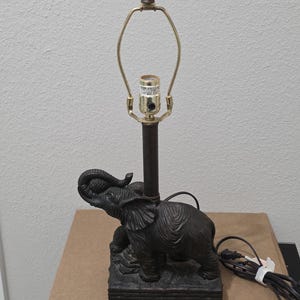 May include: A dark brown table lamp featuring a sculpted elephant base standing on a rectangular platform. The lamp includes a central metal rod, a brass-colored bulb socket with a harp attachment, and a black electrical cord with a plug.