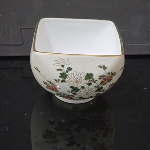 Peut inclure: Un bol carré en porcelaine blanche orné d'un motif floral délicat peint à la main avec des chrysanthèmes blancs, verts et orange. Le bord du bol est fini avec une fine bordure métallique dorée, et l'intérieur est d'un blanc uni.