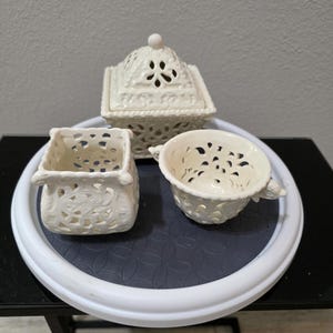 May include: Three white ceramic decorative pieces on a white and gray tray. The pieces include a small square container, a lidded box, and a small cup, all with openwork designs. The tray sits on a black table.