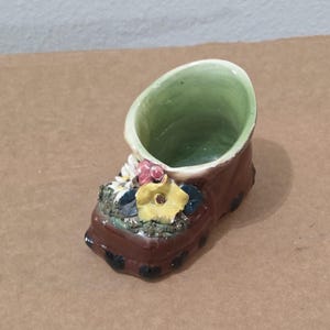 May include: A brown ceramic shoe-shaped planter featuring a glossy finish and black polka dot accents. The interior is glazed in a light green hue. The top of the shoe is decorated with a cluster of sculpted white, yellow, and red flowers.