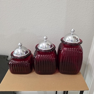 Puede incluir: Un juego de tres frascos de vidrio rojo oscuro de diferentes alturas, cada uno con una base cuadrada con detalles acanalados verticales y una tapa de metal plateado pulido extraíble con un pequeño pomo redondeado.