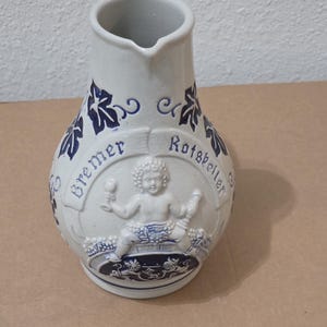 Puede incluir: Una jarra de cerámica de color crema con un diseño en relieve azul de un querubín sosteniendo una copa y un bastón. El texto "Bremer Ratskeller" está grabado en una pancarta sobre la figura, rodeado de patrones de hojas de vid azules.