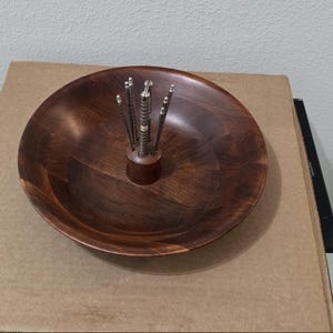 May include: A round, dark brown wooden bowl featuring a central cylindrical wooden stand. The stand holds a set of five slender, metallic cocktail picks with textured handles and small, rounded silver-toned tips arranged vertically.