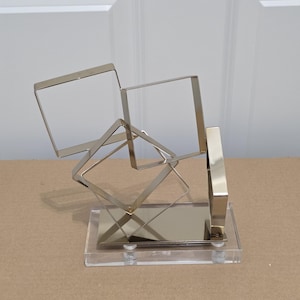 May include: A decorative sculpture featuring three interconnected metallic square frames in a polished silver finish. The frames are mounted on a rectangular clear acrylic base with a reflective silver plate, creating a geometric display piece.