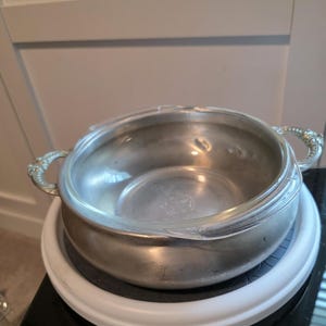May include: A silver-toned metal serving bowl with ornate handles and a clear glass lid. The bowl sits on a white and black rotating base, suggesting it's designed for easy access. The design is classic and elegant.
