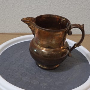 May include: A metallic pitcher with a dark bronze and copper-toned finish. The vessel features a rounded body, a wide pouring spout, and a curved handle with decorative detailing. The surface exhibits a reflective, tarnished appearance throughout.