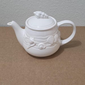 Può includere: Una teiera in ceramica bianca caratterizzata da un motivo in rilievo di varie conchiglie e linee ondulate attorno al corpo. Il coperchio è sormontato da una maniglia a forma di conchiglia scolpita, con un beccuccio curvo e un manico laterale.