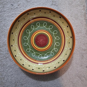 May include: A decorative ceramic bowl with a colorful, concentric design. The bowl features a dark red center, surrounded by yellow, green, and tan bands. White swirl patterns and small black dots add detail to the design. The rim is a warm orange.