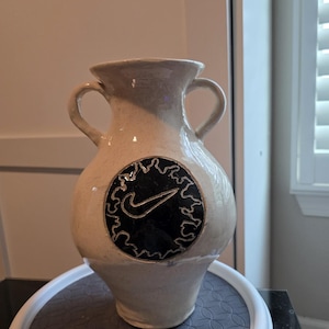 May include: A cream-colored ceramic vase with two side handles and a glossy finish. The front features a circular black medallion with a white swoosh logo surrounded by a decorative, irregular, hand-carved border design.