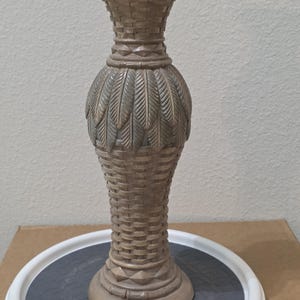 Puede incluir: Un jarrón decorativo alto de color marrón claro con un patrón de tejido de cesta texturizado en el cuello y la base, con una banda central de motivos tallados en forma de hojas superpuestas. Tiene un borde acampanado y una base circular.