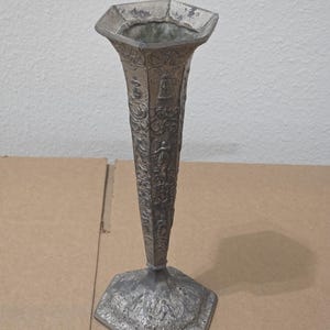 Könnte beinhalten: Eine silberfarbene, antike Vase mit sechseckigem Sockel und ausgestelltem Oberteil. Die Vase weist aufwendige geprägte Designs auf, darunter Figuren und florale Muster. Die Vase hat einen schlanken, konischen Körper und ist wahrscheinlich aus Metall.