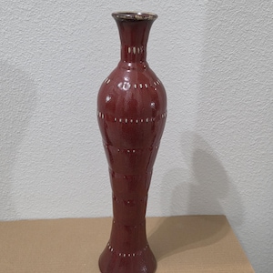 May include: A tall, slender ceramic vase with a glossy deep red glaze. The vase features a narrow neck, a bulbous midsection, and a flared base. It is decorated with horizontal bands of small, white, hand-painted dash patterns around the body.