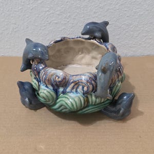 Puede incluir: Un cuenco redondo de cerámica con un patrón de olas esculpidas en tonos azules, verdes y marrones. El exterior está decorado con varias figuras de delfines grises situadas alrededor del borde y la base, creando un aspecto texturizado.
