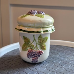 May include: A ceramic jar with a lid, featuring a grape design. The jar is white with a green rim and a grape cluster and leaves motif. The lid is topped with purple grapes. The jar is on a gray surface.