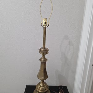 May include: A tall brass table lamp base with a polished metallic finish. The lamp features a tiered circular base, a slender central column with a decorative flared section, and a brass socket assembly with a wire harp attached at the top.