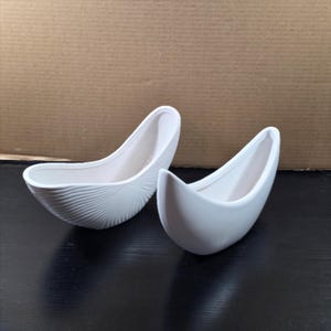 May include: Two white ceramic vessels shaped like curved boats. The vessel on the left features a textured exterior with fine, radiating lines, while the vessel on the right has a smooth, plain white surface. Both items have an open, elongated top.