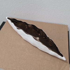 Puede incluir: Un recipiente de cerámica largo y estrecho en forma de barco con un borde irregular y ondulado. La superficie exterior tiene un acabado blanco mate, mientras que el interior presenta un esmalte de bronce metálico oscuro, liso y brillante.