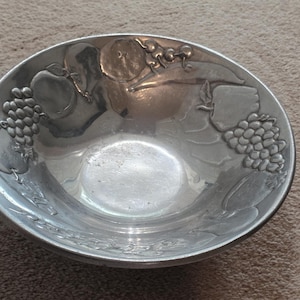 May include: A silver-colored metal bowl with embossed fruit designs, including grapes, apples, and other fruits. The bowl has a wide, shallow shape and a smooth, reflective surface. The fruit designs are raised and detailed.