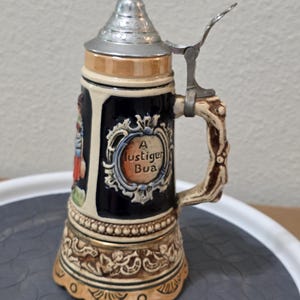 May include: A ceramic beer stein with a metallic conical lid and a hinged thumb lever. The body is dark blue with a cream-colored base and handle. It features a central oval medallion with the text A lustiger Bua and relief carvings of figures.