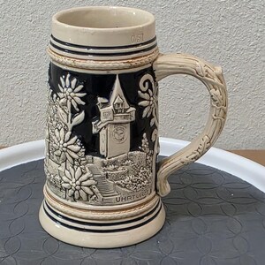 May include: A ceramic beer stein with a detailed relief design. The stein features a black background with a cream-coloured relief of a clock tower, flowers, and foliage. The handle is shaped like a tree branch, and the base is cream-coloured.