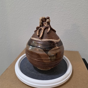 Puede incluir: Un recipiente de cerámica marrón de forma redonda con cuello cónico y una tapa escultórica texturizada. El cuerpo presenta estrías horizontales y patrones de esmalte moteado en tonos marrón, negro y crema, con un acabado brillante.