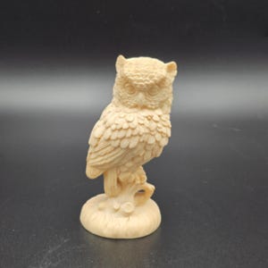 May include: A cream-colored owl figurine standing on a textured circular base. The owl features detailed carved feathers, prominent eyes, and ear tufts, with the entire sculpture rendered in a monochromatic, light beige tone with a matte finish.
