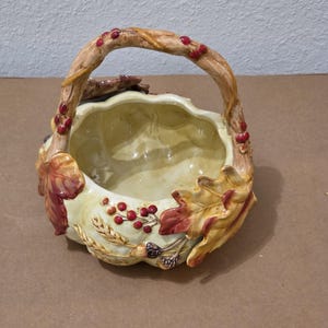 May include: A ceramic basket with a handle, decorated with autumn motifs. The cream-coloured basket has a scalloped rim and is adorned with fall leaves, wheat stalks, and red berries. The handle is designed to resemble a tree branch.