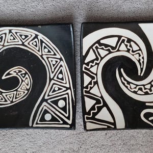 Op de afbeelding: Twee vierkante, zwarte decoratieve borden met witte tribale spiraal- en geometrische ontwerpen. De borden hebben een licht gebogen rand en worden naast elkaar weergegeven. De ontwerpen doen denken aan oude kunst.
