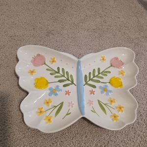 Könnte beinhalten: Weißes Keramik-Serviertablett in Schmetterlingsform mit hellblauem Mittelteil und gewellten Rändern. Das Tablett ist mit rosa, gelben und blauen Blumen und grünen Blättern verziert. Ein dekoratives Stück.
