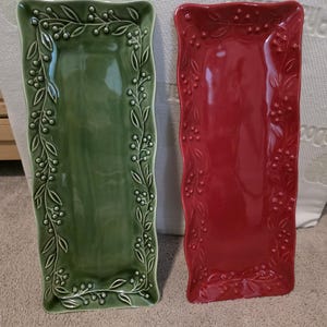 May include: Two rectangular ceramic serving platters. One is a deep green, the other is a vibrant red. Both have a raised border design of leaves and berries. The platters are ideal for serving appetizers or desserts.
