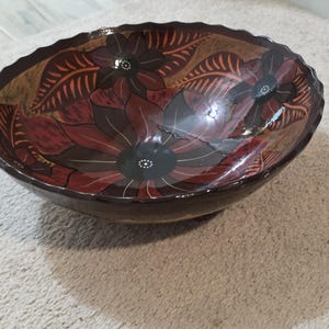 Puede incluir: Un cuenco decorativo de madera con exterior marrón oscuro y diseño floral en el interior. Las flores son en tonos rojos, marrones y negros, con patrones de hojas detallados. El cuenco tiene un borde festoneado.