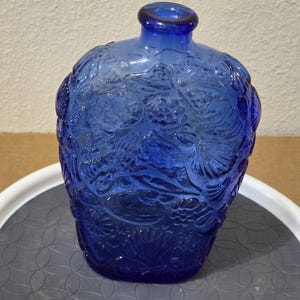 May include: A cobalt blue glass bottle featuring a raised, embossed texture of various seashells, starfish, and coral patterns across its entire surface. The bottle has a rounded, flattened body shape with a short, narrow neck and a circular opening.